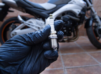 A hand with black latex gloves holds a motorcycle spark plug. A motorcycle in the background. Maintenance and spare parts concept.