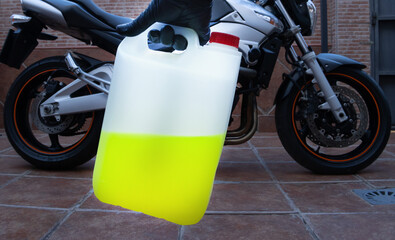 A hand with black latex gloves holds a canister of motorcycle coolant. A motorcycle in the background. Maintenance concept.