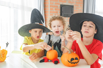 Fototapeta premium Three real siblings two brothers and sister making together jack o lantern at their home white kitchen, cutting real pumpkins and playing with paper black ghosts, lovely vibe of halloween