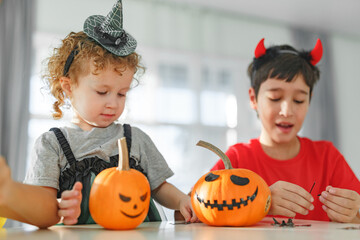 Two real happy siblings brother and sister cutting together from real natural pumpkin jack o lantern, while playing with paper decorations of ghosts, children wearing with witch hats, halloween
