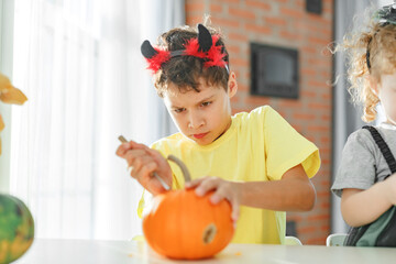 Two real happy siblings brother and sister cutting together from real natural pumpkin jack o lantern, while playing with paper decorations of ghosts, children wearing with halloween costumes
