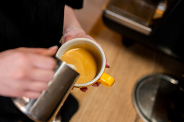 Close-up of hands pouring milk into a yellow cup with espresso to create latte art, against a blurred background. Perfect for coffee shop promotions, barista training, or beverage-related content.