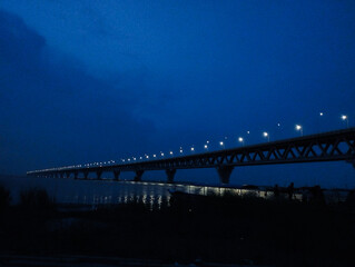 Fototapeta premium Night Vision of the Padma Bridge. A serene night view of the Padma Bridge, illuminated by rows of streetlights that cast reflections on the calm waters below. The bridge stretches across the horizon, 