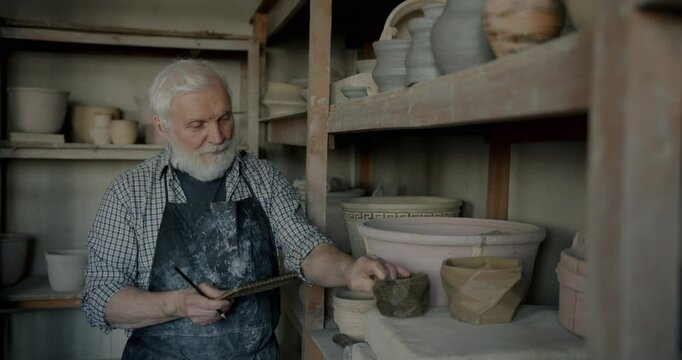 Portrait of senior man pottery business owner checking pots on shelves doing inventory developing studio. People and entrepreneurship concept.