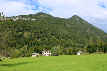 Vallata con prato di erba tre case boschi montagne e cielo con nuvole in Val di Sella