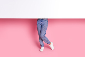 Cropped photo of woman in stylish denim jeans and white stylish sneakers hiding behind whiteboard isolated on pink color background