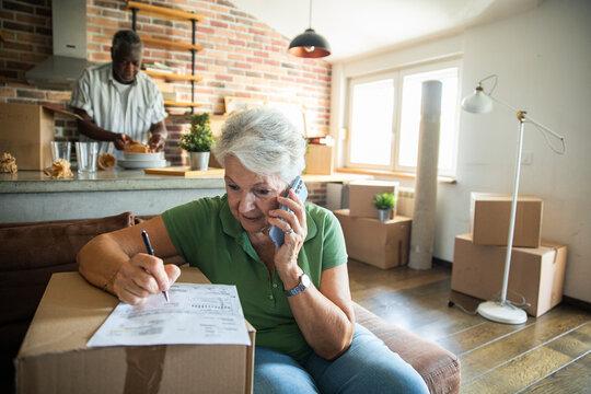 Senior woman making a phone call while reviewing paperwork in living room with moving boxes