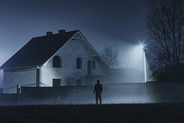A man stands in front of a house at night. A silhouette of a man under a street lamp, exuding mystery on a foggy night. A lone figure walks toward a dimly lit house on a foggy night. © Oleksandr