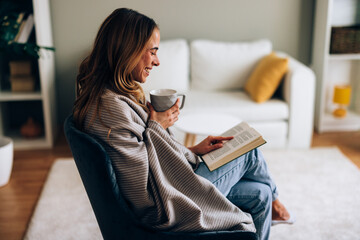 Young woman enjoying a quiet moment at home wrapped in a blanket reading a book and sipping tea
