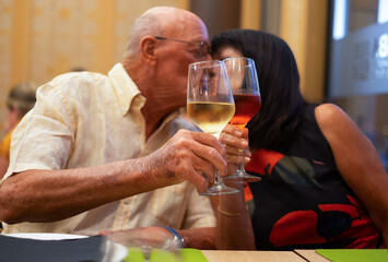 Senior caucasian couple sitting at restaurant table kissing and toasting with two glasses of wine, red and white