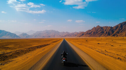 Naklejka premium A motorcyclist rides along a vast, open road surrounded by dramatic mountains and a clear blue sky in a desert landscape.
