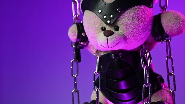 A teddy bear in a mask is suspended from chains in leather handcuffs and accessories for bdsm games and sex in neon light against a background of copy paste