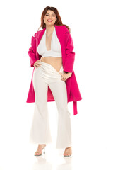 Fashion style studio portrait of beautiful young woman. White wide leg pants and pink coat. Model sitting and standing against white background