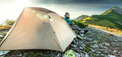 Bearded man wearing down jacket sits beside his tent in front of a mountain peak grinding coffee © XArt