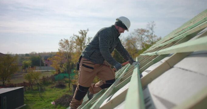 Construction worker hammering roof beams on building structure, wearing safety helmet, high angle view of teamwork with colleagues in background, outdoor construction project site