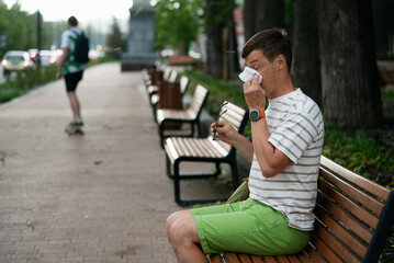Obraz premium Man is blowing his nose with tissue while sitting on bench in the park, experiencing symptoms of cold, flu, or allergy, holding eyeglasses in his hand
