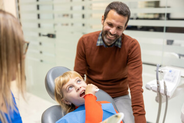 Obraz premium A child sits in a dentist chair, wearing a dental bib, while a smiling adult stands beside them in a modern dental office, suggesting a comforting environment.