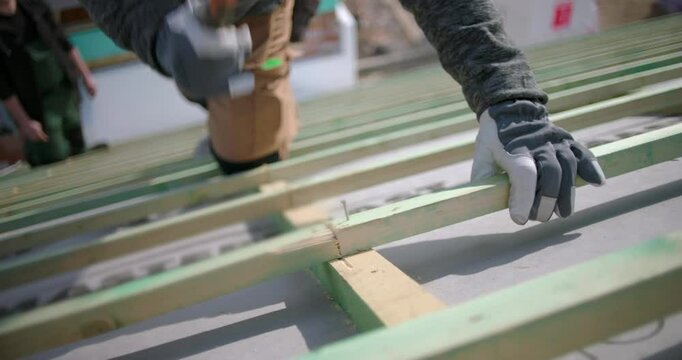 Close-up of construction worker hammering nails into roof beams, wearing safety gloves, focus on hands and hammer, outdoor construction project with colleagues in background