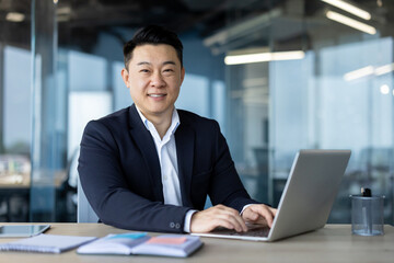 Portrait of a smiling young Asian male businessman sitting at the workplace with a laptop, looking confidently at the camera