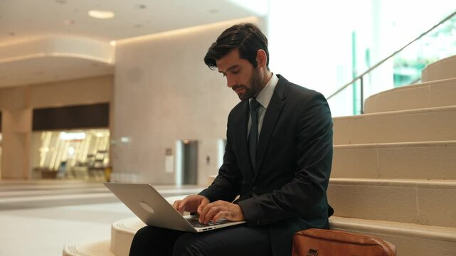 Skilled investor working or planing strategy by using laptop at stair. Business man wearing suit while working and sitting at stairs. Smart manager finish working and going to another place. Exultant.