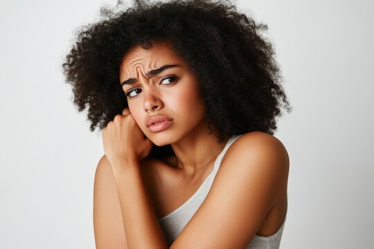 Woman experiencing shoulder pain, indoor, light grey background, standing with one hand gripping her shoulder.