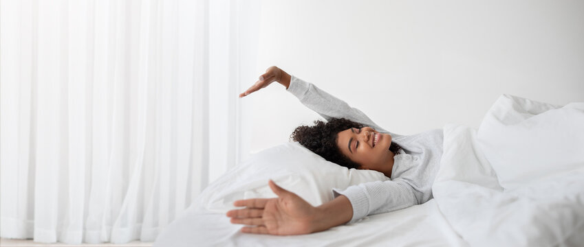 African American woman wakes up and stretches her arms wide while lying on a comfortable bed. Sunlight filters through the curtains, creating a warm atmosphere in her serene bedroom.