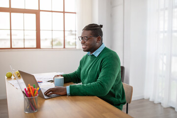 A black guy wearing a green sweater and glasses is working diligently on a laptop at a sunny desk....