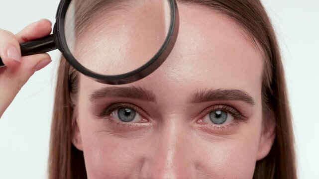 Blonde woman with blue eyes on green background holding magnifying glass and showing her wrinkles on face
