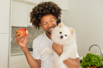 A black man smiles broadly in a bright kitchen, holding an apple in one hand and a fluffy white dog in the other. Fresh produce is visible on the counter, adding to the cheerful atmosphere.