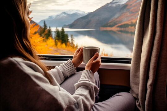 Mujer desayunando caf&eacute; caliente dentro de una casa m&oacute;vil con vistas a un paisaje de monta&ntilde;as y lago en oto&ntilde;o. Estilo de vida de viaje con camper.