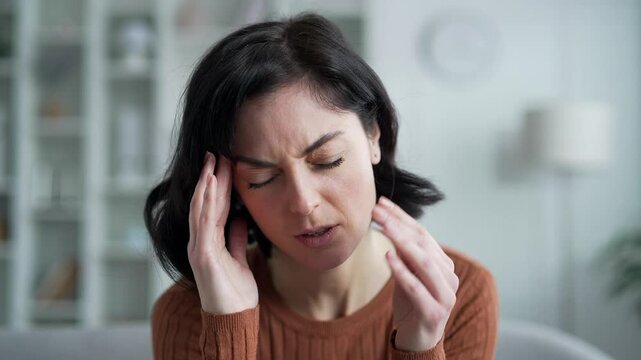 Young adult woman suffering from headache while sitting on sofa in living room at home. Upset female massages her temples with her hands. She is tired, feels pain, stress and fatigue. Close up