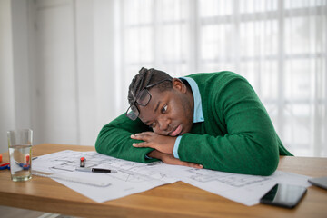 A young black man with glasses appears tired and frustrated while resting his head on a cluttered...