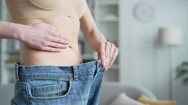 Young adult woman displaying weight loss achievement wearing oversized jeans standing in living room at home. Concept of fitness, personal achievement, health transformation, lifestyle change Close up