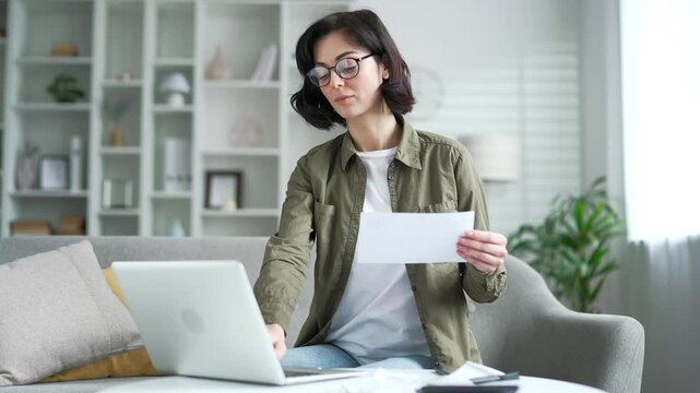Young adult woman dealing with household finances using website or app on laptop sitting on couch at home office. The female holds a utility bill and forms payments online by entering data on computer
