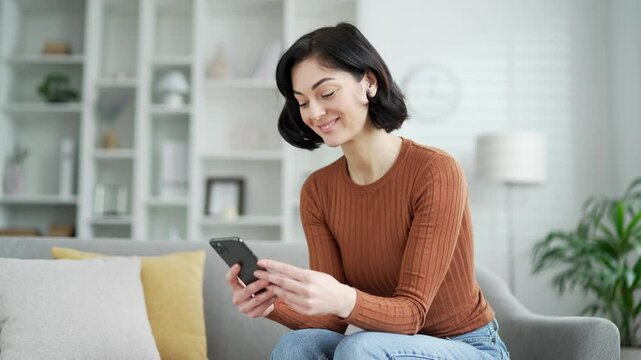 Young adult woman in cozy living room sitting on sofa, smiling at phone, enjoying casual time at home. Smiling female student browses social networks, makes purchases, chats, writes or reads messages - Powered by Adobe