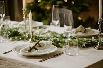 Medium close up of snow white table with cotton napkin in napkin ring surrounded with polished crystal glasses and pine garland
