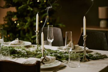 Medium close up extinguished candles in silver candlesticks on table decorated with pine garlands and polished glasses for champagne