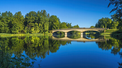 Fototapeta premium A beautiful bridge arches over a still lake, its reflection shimmering in the water. The Rakotz bridge in Kromlau, Germany, is a stunning sight, framed by lush trees and a clear blue sky.