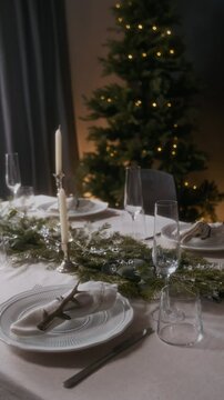 Vertical shot of holiday table with elegant white plates, candles and evergreen branch centerpiece in cozy room decorated with Christmas tree