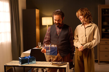 Mature Jewish man with yarmulke on head lighting candle with help of other one to keep Hanukkah tradition, his blonde Caucasian wife looking at process and smiling