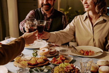 Medium close up of unrecognizable friends clinking crystal glasses with red wine while celebrating seasonal holiday at table with different types of treats