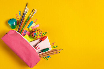 Top view a bright collection of school supplies, including pens, pencils, paper clips, and a sharpener in a pink pencil case, set against a vibrant yellow background