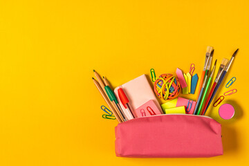 Top view a bright collection of school supplies, including pens, pencils, paper clips, and a sharpener in a pink pencil case, set against a vibrant yellow background