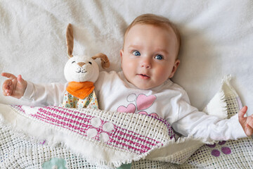 A smiling baby lies in bed under a blanket, holding a plush toy rabbit. The scene radiates warmth...