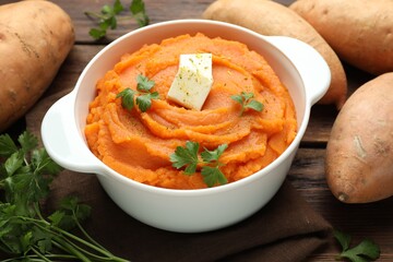 Tasty mashed sweet potato with butter in bowl, fresh vegetables and parsley on wooden table, closeup