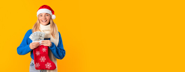 Happy woman wearing knitted sweater and Santa hat, holding Christmas sock with fan of cash money in...