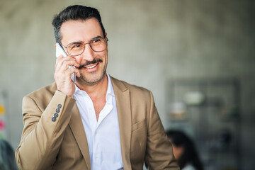 A man in a tan blazer is smiling while on a phone call in a contemporary office. The background features a blurred workspace, indicating a professional environment.