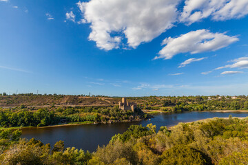 A historic castle stands majestically atop a rocky island surrounded by calm river waters, with a backdrop of lush hills