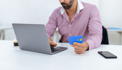 A man sits at a white office desk, focused on his laptop while holding a credit card in his right hand. A cup of coffee and a smartphone rest nearby, highlighting a work setting.