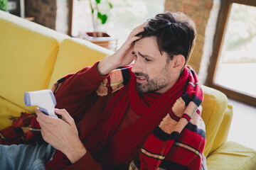 Young man feeling unwell indoors, wrapped in a colorful blanket, checking temperature on a digital...